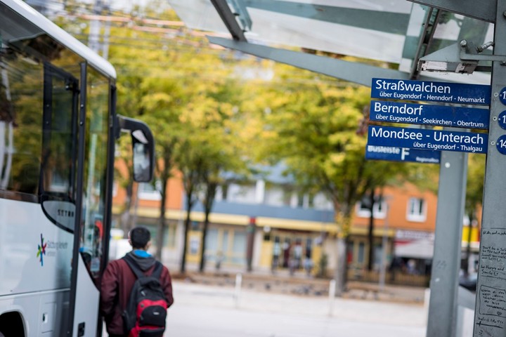 Passenger boarding a regional bus at a bus stop, signposts to destinations in the Salzburg area in the background.