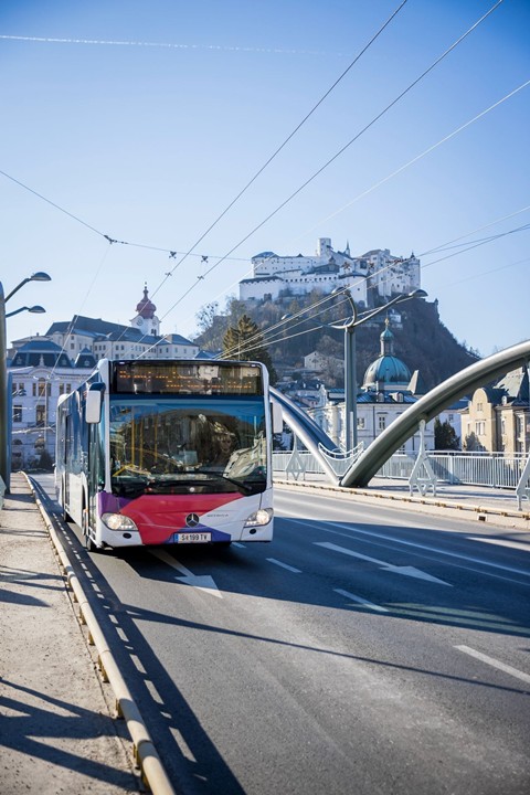 A public bus drives over the State Bridge in Salzburg, with Hohensalzburg Fortress above the old town in the background.